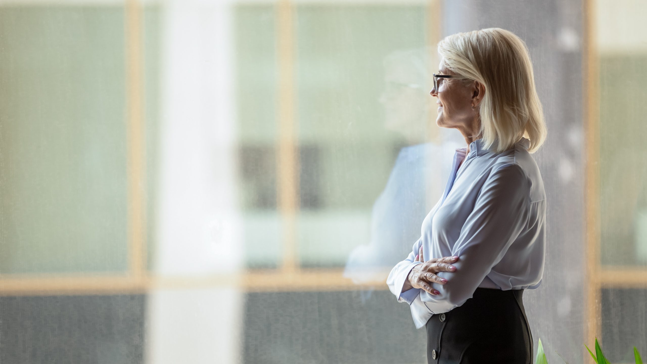 Happy mature Caucasian leader stand at window in office look in distance thinking or visualizing, smiling successful middle-aged woman boss dream of career perspective, business vision concept